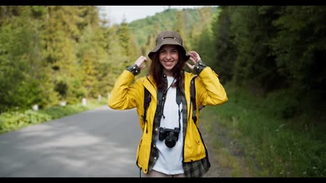 Portrait-of-a-tourist-girl-in-special-hiking-clothes,-along-with-a-camera-in-a-yellow-jacket,-who-looks-into-the-camera-against-the-backdrop-of-a-mountain-forest