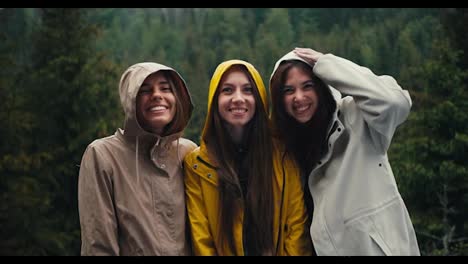 Three-girls-in-light-jackets-in-hoods-rejoice-in-the-coming-rain-and-look-at-the-camera-and-hug-in-a-mountain-forest