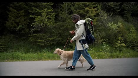 A-man-of-tourists-in-special-clothes-for-a-hike-with-a-large-backpack-with-his-light-colored-dog-walks-along-the-forest-road
