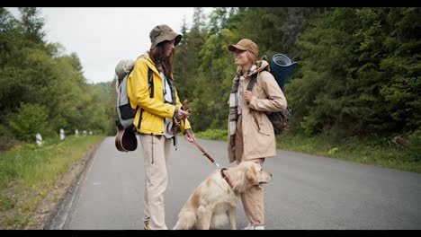 2-girls-on-a-hike.-A-brunette-girl-in-a-yellow-jacket-holds-her-light-colored-dog-on-a-leash-while-chatting-with-her-friend-near-the-road-in-the-middle-of-the-forest