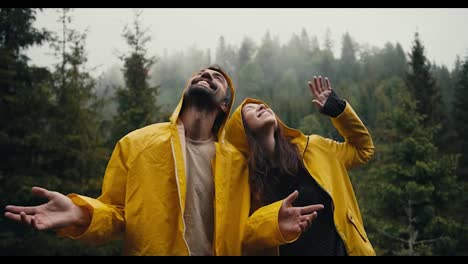 Happy-couple:-a-guy-and-a-girl-in-yellow-jackets-look-at-the-sky,-smile-and-wait-for-the-coming-rain-against-the-backdrop-of-a-mountain-coniferous-forest