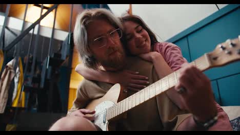 A-blonde-with-long-hair-in-glasses-with-a-beard-plays-the-electric-guitar,-a-brunette-girl-hugs-his-shoulders-and-listens-to-the-guy-playing-in-a-cozy-room