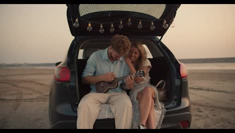 A-bearded-man-with-curly-hair-in-a-blue-shirt-is-playing-the-ukulele-and-his-blonde-girlfriend-is-hugging-him,-they-are-laughing-and-sitting-in-the-trunk-of-the-black-car-decorated-with-the-lights-against-the-yellow-sky,-while-their-dog-is-lying-on-the-sand