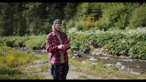A-young-male-tourist,-blond-in-glasses-with-a-beard-in-a-red-hiking-shirt,-unpacks-the-tent.-The-guy-opens-the-frame-of-the-tent-and-tries-to-fold-it-into-a-single-structure-against-the-backdrop-of-a-forest-river-in-the-mountains