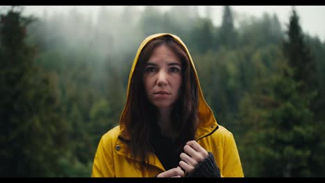 A-sad-girl-in-a-yellow-jacket-with-a-hood-stands-against-the-backdrop-of-a-coniferous-mountain-forest,-she-is-sad-and-looks-at-the-camera