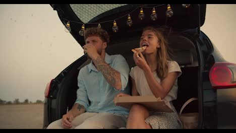 A-bearded-man-with-curly-hair-in-a-blue-shirt-is-drinking-tea-next-to-his-blonde-girlfriend,-who-is-eating-pizza,-they-are-sitting-in-the-trunk-of-the-black-car-decorated-with-the-lights-against-the-background-of-the-flying-birds-in-the-yellow-sky