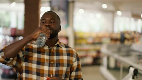 Portrait-of-a-man-with-black-skin-color-in-a-plaid-shirt-drinks-water-from-a-transparent-plastic-bottle-in-a-supermarket.-Shopping-break