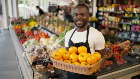 Portrait-of-a-Black-skinned-man-in-a-brown-T-shirt-and-black-apron-posing-with-a-box-of-yellow-tomatoes-in-his-hands-in-a-supermarket