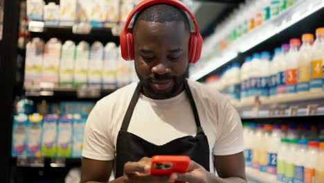 a-man-with-Black-skin-color-in-red-headphones-and-a-black-apron-with-a-red-smartphone-walks-along-the-dairy-department-in-a-supermarket