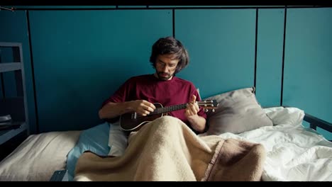 Ein-Brünetter-Mann-Mit-Bart,-Rotem-Hemd-Und-Brille-Spielt-Gitarre-Und-Liegt-Auf-Einem-Bett-Mit-Blauen-Wänden