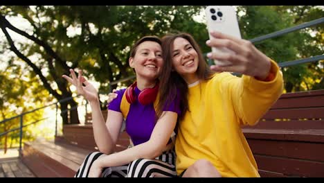 A-girl-with-a-short-haircut-in-a-purple-top-and-a-girl-in-a-yellow-sweater-take-a-selfie-using-a-white-phone-in-the-park-in-summer
