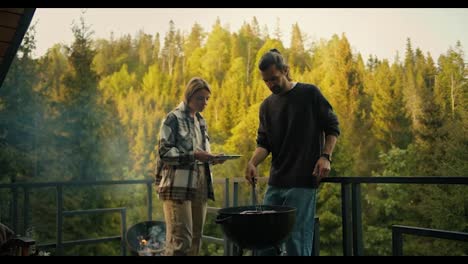 A-blonde-girl-helps-a-brunette-guy-cook-meat-on-the-grill-on-the-balcony-of-a-country-house-against-the-backdrop-of-a-coniferous-forest