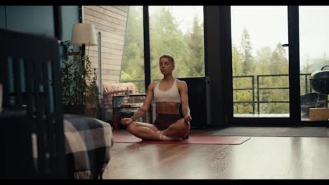 A-blonde-girl-in-a-white-top-with-tattoos-is-engaged-in-meditation-and-yoga-on-a-special-rug-in-a-modern-house-against-the-backdrop-of-a-green-forest-outside-the-window
