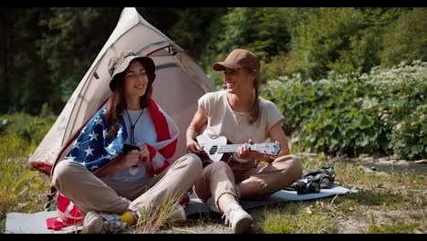 A-brunette-girl-is-wrapped-in-the-flag-of-the-United-States-of-America-and-a-girl-in-camping-clothes-plays-the-guitar,-they-are-sitting-near-the-tent-against-the-backdrop-of-a-green-forest