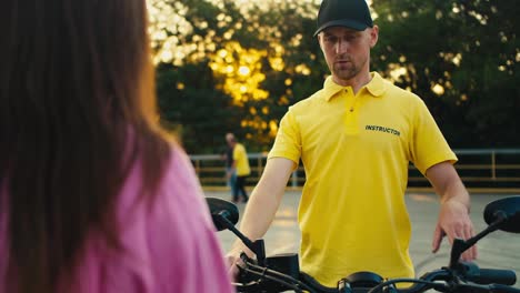 A-guy-in-a-cap-in-a-yellow-t-shirt-of-an-instructor-sits-on-a-motorcycle-and-shows-a-girl-in-a-pink-t-shirt-How-to-sit-on-a-motorcycle.-driving-school-instructor