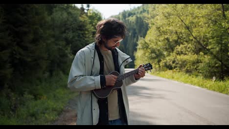 A-brunette-guy-in-a-white-jacket-near-the-road-plays-a-musical-stringed-instrument-on-a-hike-in-the-mountains