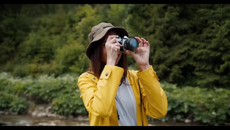 A-brunette-girl-in-a-yellow-hiking-jacket-photographs-a-forest-and-a-mountain-river-with-a-camera