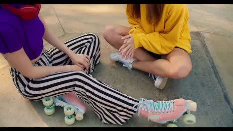 Close-up-shot-of-a-girl-in-a-yellow-sweater-and-a-girl-in-a-purple-top-and-striped-pants-in-pink-roller-skates-sit-on-a-concrete-floor-in-a-skate-park-in-summer
