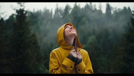 A-happy-girl-in-a-yellow-jacket-smiles-and-waits-for-the-coming-rain-against-the-backdrop-of-a-green-coniferous-forest-in-the-mountains