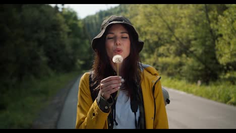 Happy-brunette-girl-tourist-in-special-clothes-for-hiking-in-a-yellow-jacket-blows-on-a-dandelion-that-is-flying-against-the-background-of-the-forest
