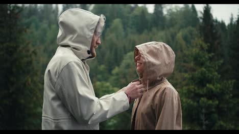 The-guy-fastens-the-girl's-jacket-and-kisses-her-during-the-rain-in-a-mountain-coniferous-forest.-Happy-couple-against-green-forest-background