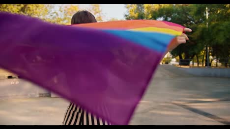 Portrait-of-a-happy-girl-in-a-purple-top-and-striped-pants-who-rides-on-roller-skates-in-a-skate-park-and-holds-an-LGBT-flag-that-flutters-in-the-wind