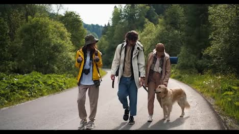 A-guy-and-two-girls-in-special-clothes-for-a-hike-along-with-their-light-colored-dog-walk-along-the-road-along-the-forest-in-a-mountainous-area
