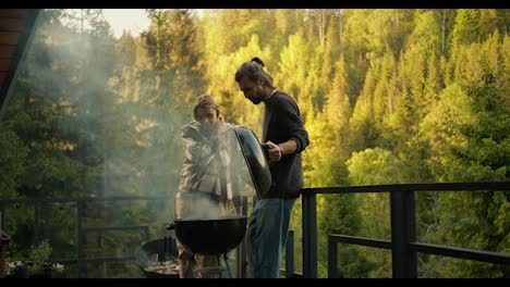 A-brunette-guy-and-a-blonde-girl-are-watching-the-preparation-of-meat-on-the-grill-during-a-picnic-on-the-terrace-against-the-backdrop-of-a-coniferous-forest-and-mountains