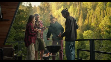 A-group-of-friends-are-relaxing-and-grilling-meat-on-the-terrace-of-a-country-house-against-the-backdrop-of-a-green-coniferous-forest.-The-guy-plays-the-guitar,-a-couple-of-people-work-with-the-barbecue