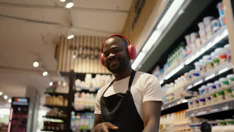 Close-up-shot-of-a-Black-skinned-man-in-red-headphones-and-a-black-apron-dancing-in-the-dairy-department-of-a-supermarket