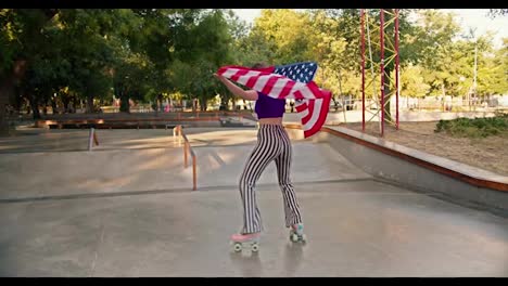 Ein-Mädchen-Mit-Einem-Kurzen-Haarschnitt,-Einem-Lila-Oberteil-Und-Einer-Gestreiften-Hose-Läuft-Im-Sommer-Rollschuh-Und-Trägt-Die-US-Flagge-In-Einem-Skatepark