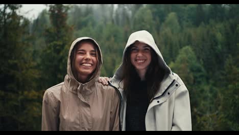 Two-girls-in-light-jackets-in-hoods-rejoice-in-the-coming-rain-and-look-at-the-camera-in-a-mountain-forest