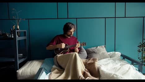 A-brunette-man-in-a-dark-red-shirt-lies-on-a-sofa-wrapped-in-a-blanket-and-plays-the-guitar-against-a-turquoise-wall