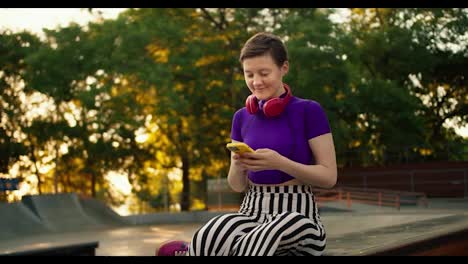 A-young-girl-with-a-short-haircut-in-a-purple-top-sits-on-social-networks-and-takes-a-selfie-using-a-yellow-phone-in-a-park-in-summer.-Teenager-in-red-headphones-in-the-park