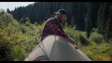 A-blond-man-in-a-red-plaid-shirt-set-up-a-tent-in-a-clearing-near-the-mountains