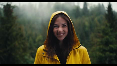 A-happy-girl-in-a-yellow-jacket-with-a-hood-stands-against-the-backdrop-of-a-coniferous-mountain-forest,-she-smiles-and-looks-at-the-camera