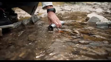 Close-up-shot:-a-guy-collects-water-in-a-thermos-from-a-mountain-river-during-a-hike