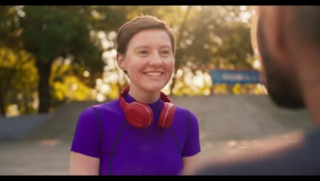 First-person-view:-A-brunette-guy-in-a-gray-t-shirt-communicates-with-a-girl-with-a-short-haircut-in-a-purple-top-and-red-headphones-in-a-skate-park-in-summer