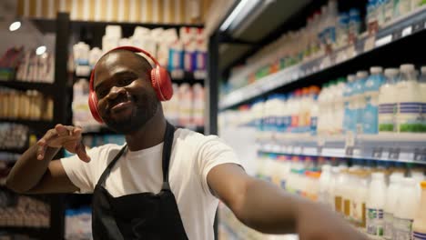 a-man-with-Black-skin-color-in-red-headphones-and-a-black-apron-dances-in-the-dairy-department-of-a-supermarket.-Happy-at-work