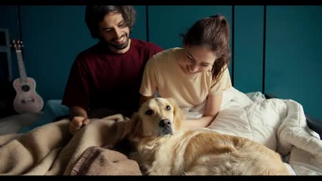 Happy-couple-together-with-their-pet:-A-guy-and-a-girl-play-with-their-dog-of-light-coloring-in-the-bed-of-their-house-against-the-background-of-a-turquoise-wall