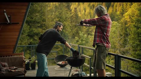 A-blond-man-and-a-brunette-man-cook-food-on-the-grill.-The-guy-in-the-red-plaid-shirt-raises-the-grill-so-that-the-guy-in-the-green-sweatshirt-pours-hot-coals.-Picnic-with-a-view-of-the-mountains-and-coniferous-forest