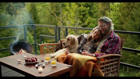 A-romantic-evening-in-a-country-house:-a-blond-man-and-a-blond-girl-are-sitting-on-a-couch-wrapped-in-a-blanket.-Near-them-sits-their-light-colored-dog.-Relax-with-a-view-of-the-mountains-and-the-forest
