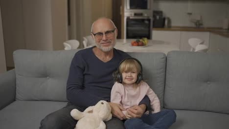 Portrait-of-a-small-girl-in-headphones-with-her-grandpa-while-sitting-on-sofa