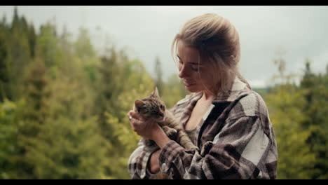 Close-up-shot:-a-happy-blonde-girl-holds-her-cat-in-her-arms-and-strokes-her-on-the-balcony-against-the-backdrop-of-a-coniferous-forest-in-the-mountains