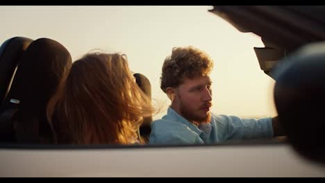A-bearded-guy-with-curly-hair-in-a-blue-shirt-drives-a-white-convertible-together-with-a-blonde-girl-and-against-the-yellow-sky
