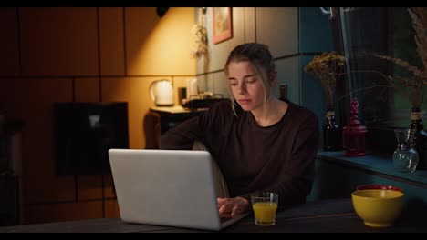 A-happy-blonde-girl-rejoices-and-looks-at-the-screen-of-a-white-laptop-in-a-dark-evening-room-illuminated-by-a-yellow-lamp