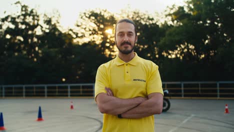 A-male-driving-instructor-in-a-yellow-t-shirt-with-a-beard-and-mustache-folded-his-arms-over-his-chest-and-poses