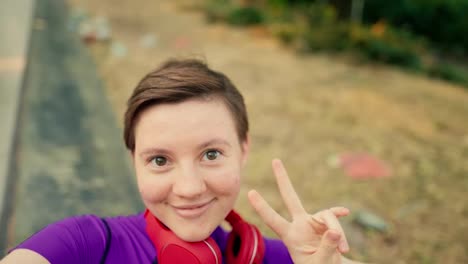 First-person-view:-A-happy-girl-with-a-short-haircut-with-brown-eyes-in-a-purple-top-and-red-headphones-lies-down-on-a-bench-and-takes-a-selfie-in-a-skatepark-in-summer
