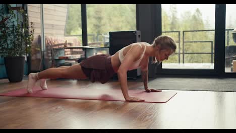 A-blonde-girl-with-tattoos-in-a-white-top-does-push-ups-on-a-special-rug-on-the-floor-of-a-country-house-overlooking-a-green-coniferous-forest