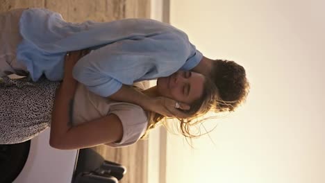 Vertical-video:-A-girl-in-a-white-top-hugs-her-bearded-boyfriend-with-curly-hair.-A-romantic-date-in-windy-weather-against-the-white-convertible,-a-river-and-a-yellow-sky